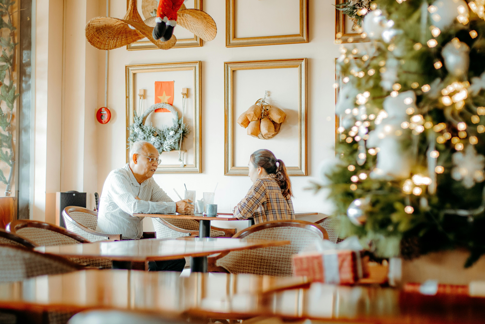 An elderly asian couple sits together at a dining table in a retirement community dining room, with a lit Christmas tree in the foreground.