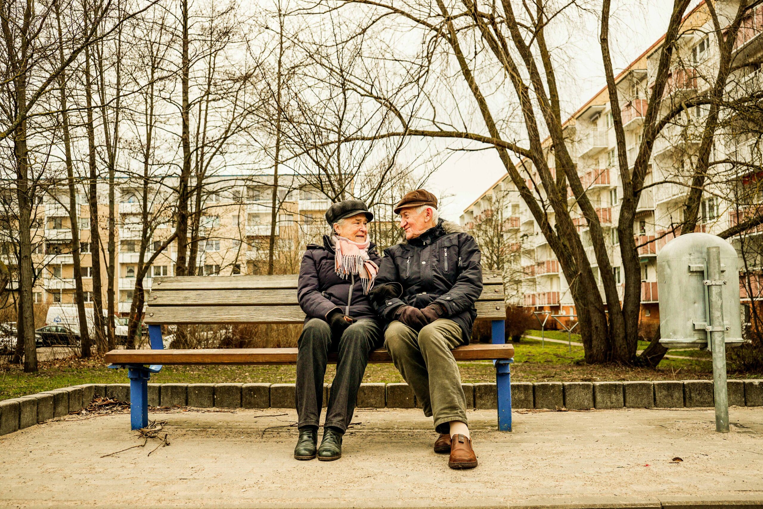 Elderly couple sitting close together on a park bench, smiling at each other on an overcast day with apartment buildings in the background.